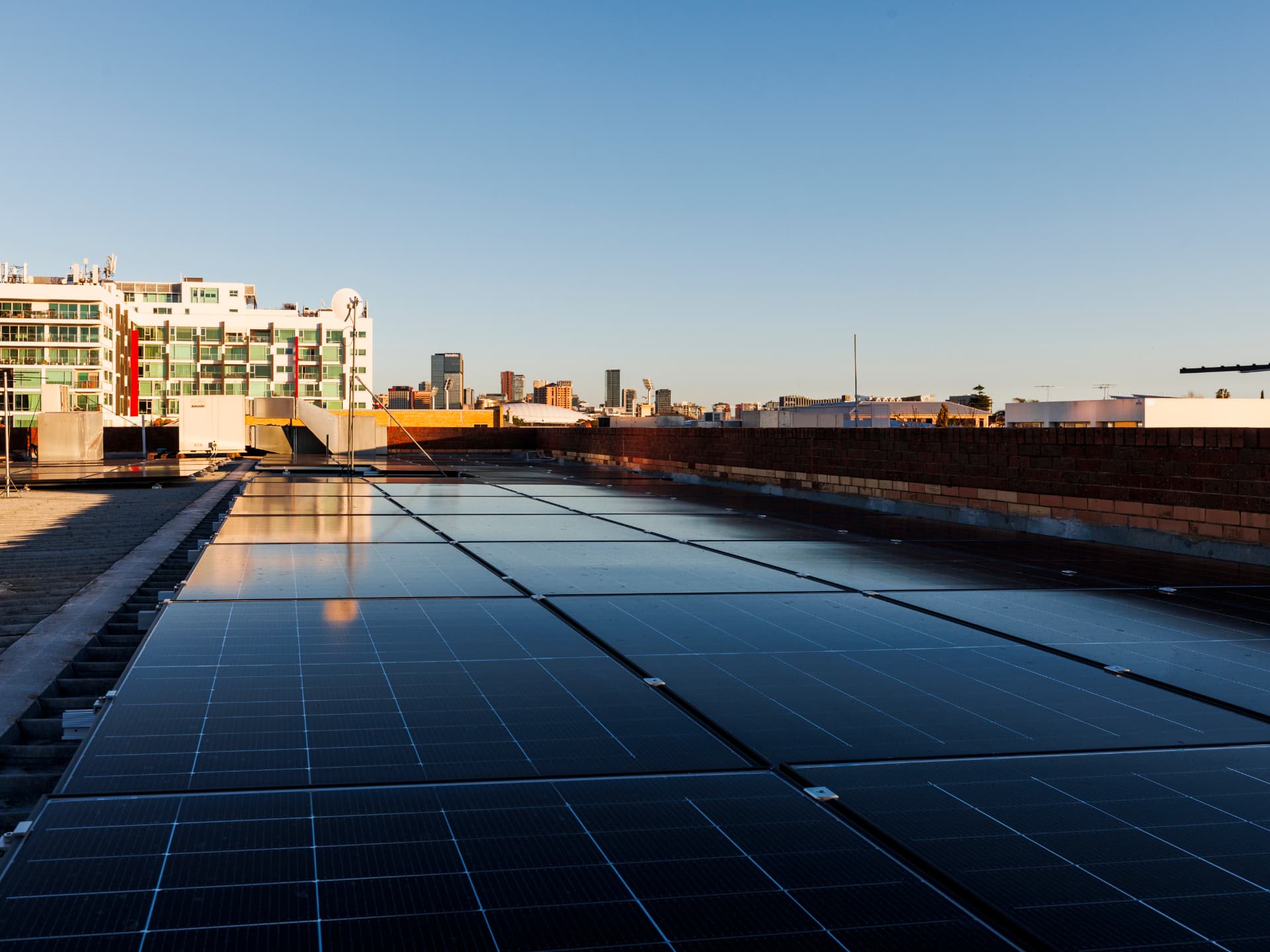 Rows of solar panels on a flat commercial rooftop at sunset, with the Adelaide city skyline in the distance