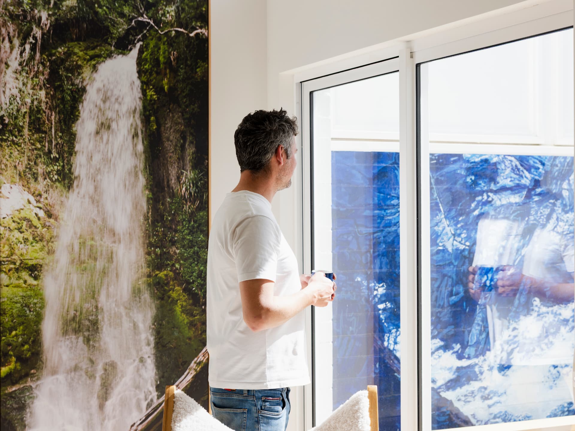 Man in a white t-shirt holding a mug, looking out through a sunlit sliding door in a home interior